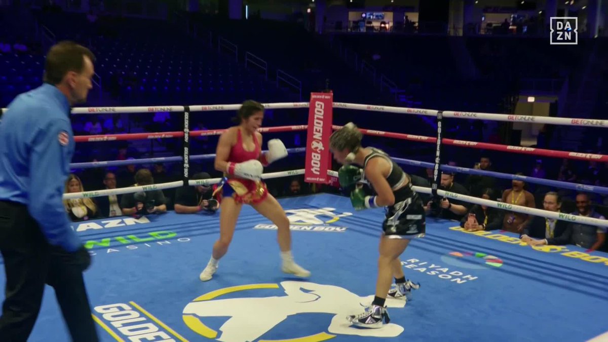 Two female boxers exchange heavy blows in a Golden Boy boxing ring on the DAZN-broadcasted Schofield vs. Bahdi undercard, with the "GOLDENBOY" and "GOLDEN" logos visible on the ring mat and ropes as a referee in blue oversees the action.