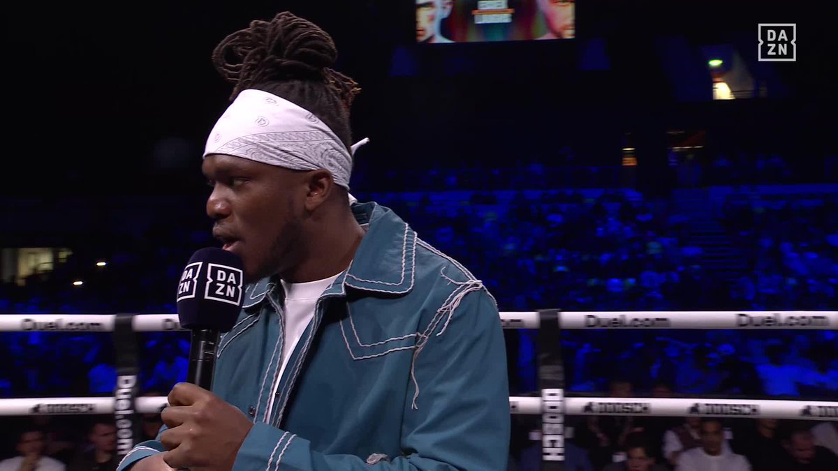 A man with a white bandana and teal jacket speaks into a "DAZN" branded microphone inside a boxing ring, likely during the announcement mentioned in the post. The "DAZN" logo appears prominently on the microphone, with "DAZN.com" and "BOXXER" branding visible on the ring ropes behind him.