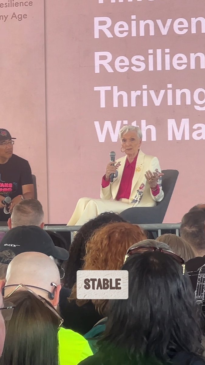An older woman with short gray hair in a yellow blazer and pink top speaking into a microphone at an event, with a banner behind her reading "Reinventing Resilience / Thriving With..."; a label in the foreground says "STABLE".