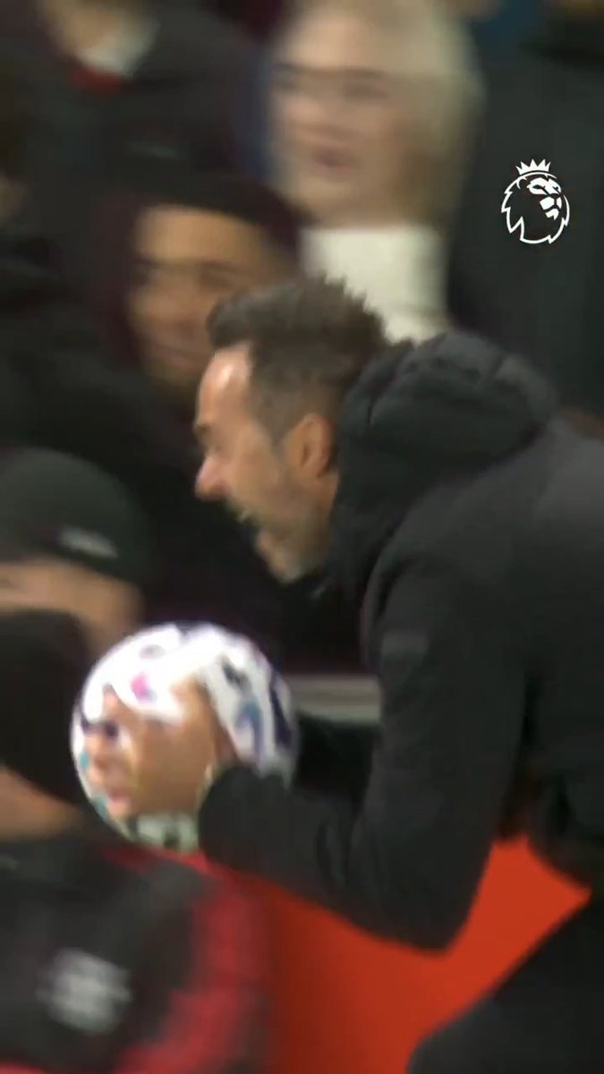 A bearded man in a black hooded jacket holds a Premier League football, appearing to prepare for a unique celebration while watching from the sidelines. The Premier League lion logo is visible in the upper right, and the man's distinctive appearance matches that of Brighton manager Roberto De Zerbi, who is known for his passionate touchline celebrations.