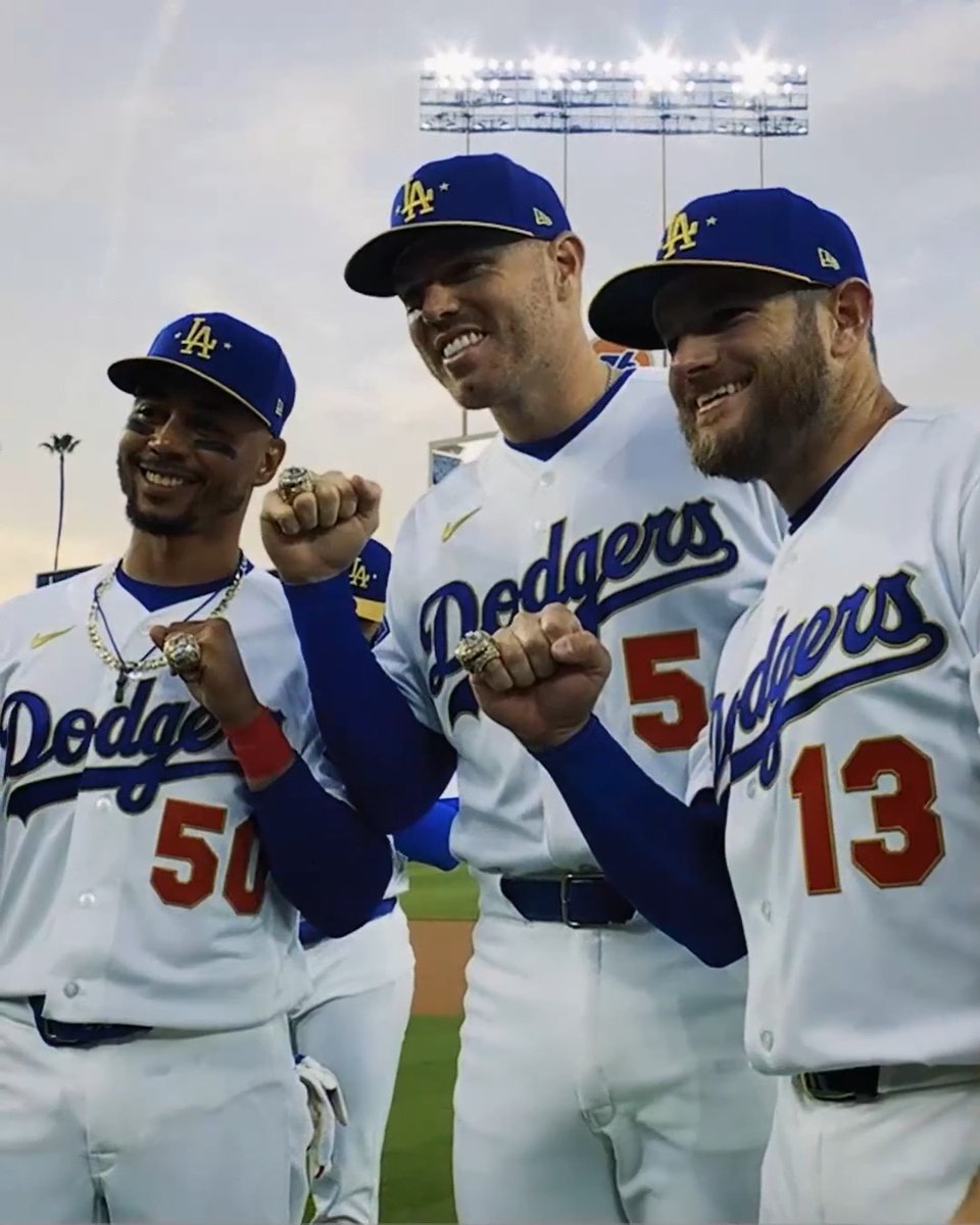 Three Dodgers players in white "Dodgers" jerseys with numbers 50, 5, and 13 proudly display their championship rings while smiling on the field. The image celebrates the team's return to the NLCS for the 5th time in 7 seasons, showcasing the winning tradition behind their postseason success.