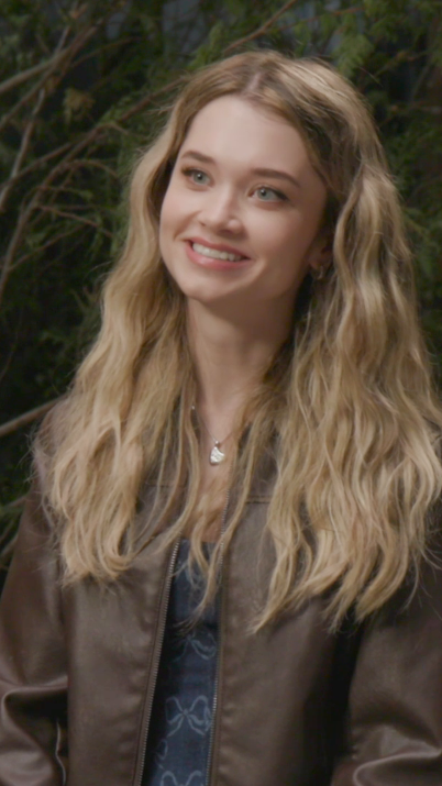 A young woman with long wavy blonde hair smiles warmly, wearing a brown leather jacket and a delicate pendant necklace, against a lush green foliage backdrop. The image appears to promote the Prime Video series "#Carrie," inviting viewers to get to know the cast.