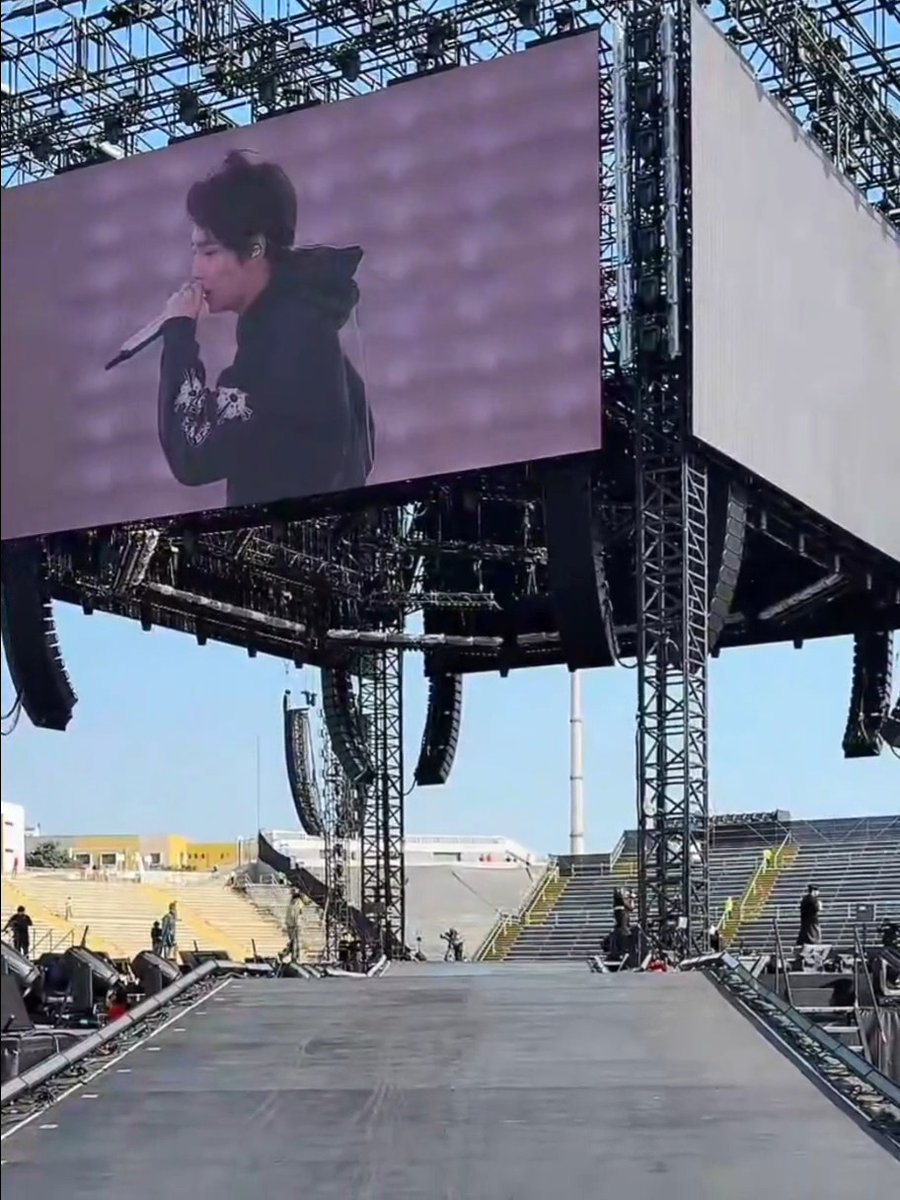 A large outdoor concert stage setup with an extended runway/catwalk, massive LED screens displaying a male singer performing with a microphone, with crew members working on the empty stadium stage below.