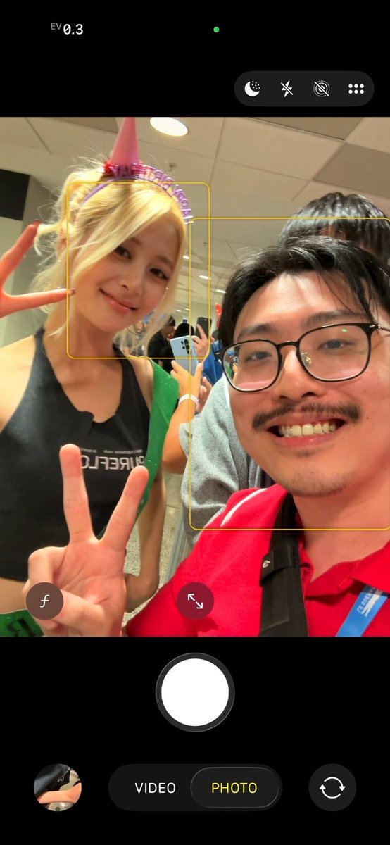 A smartphone camera interface showing a selfie of a young man in a red shirt with glasses and a young blonde woman wearing a party hat, both making peace signs.