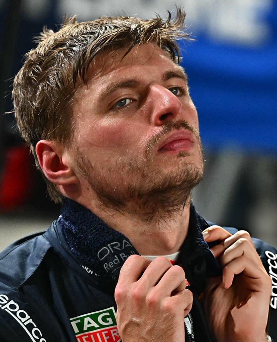 A close-up of a man with short brown hair and a beard, wearing a dark jacket with sponsor logos including 'Sparco' and 'TAG Heuer', looking to the side.