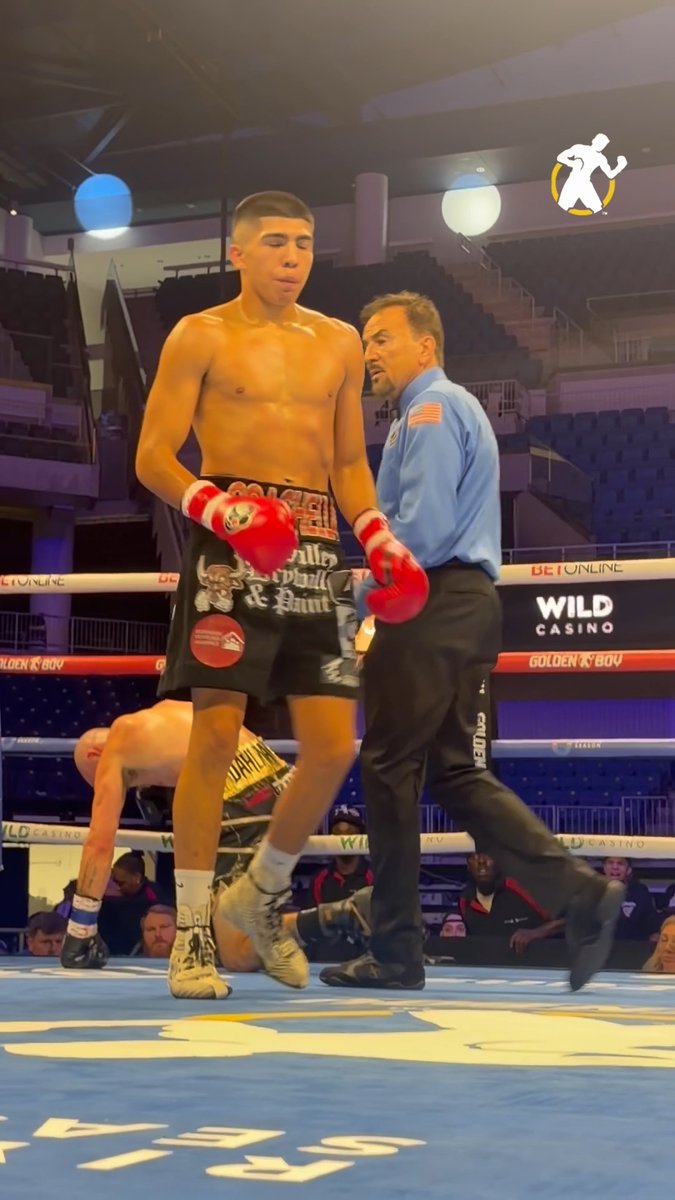 A boxer with his gloves raised walks across the ring with a referee wearing a blue shirt with an American flag patch, while another fighter is down on the mat behind them, capturing the moment of a 2nd round TKO victory for Grant Flores as advertised on the DAZN broadcast.