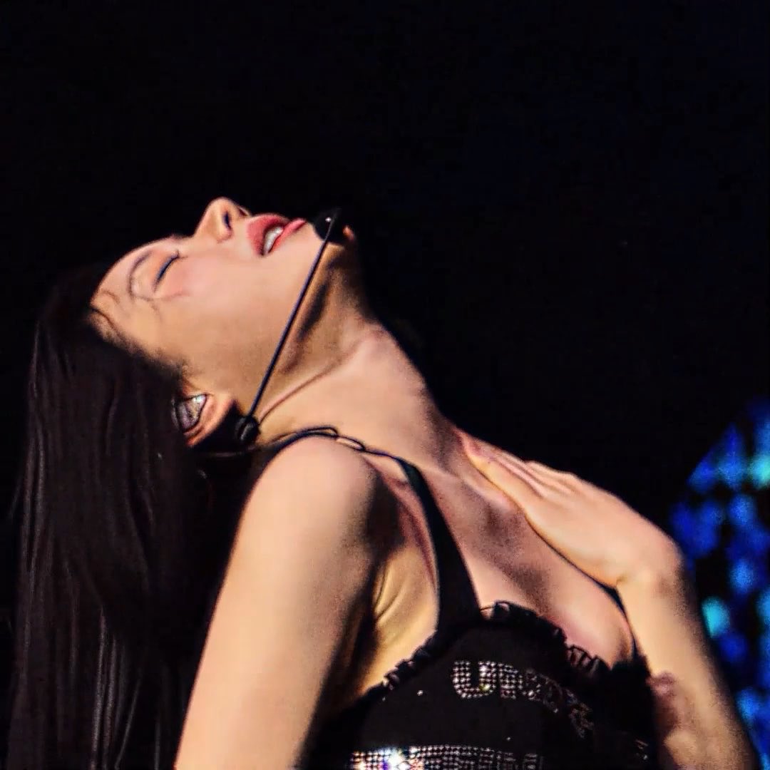 A close-up performance photo of a woman with long dark hair wearing a headset microphone and a sequined top, head tilted back in a dramatic pose against a dark background with blue stage lighting.