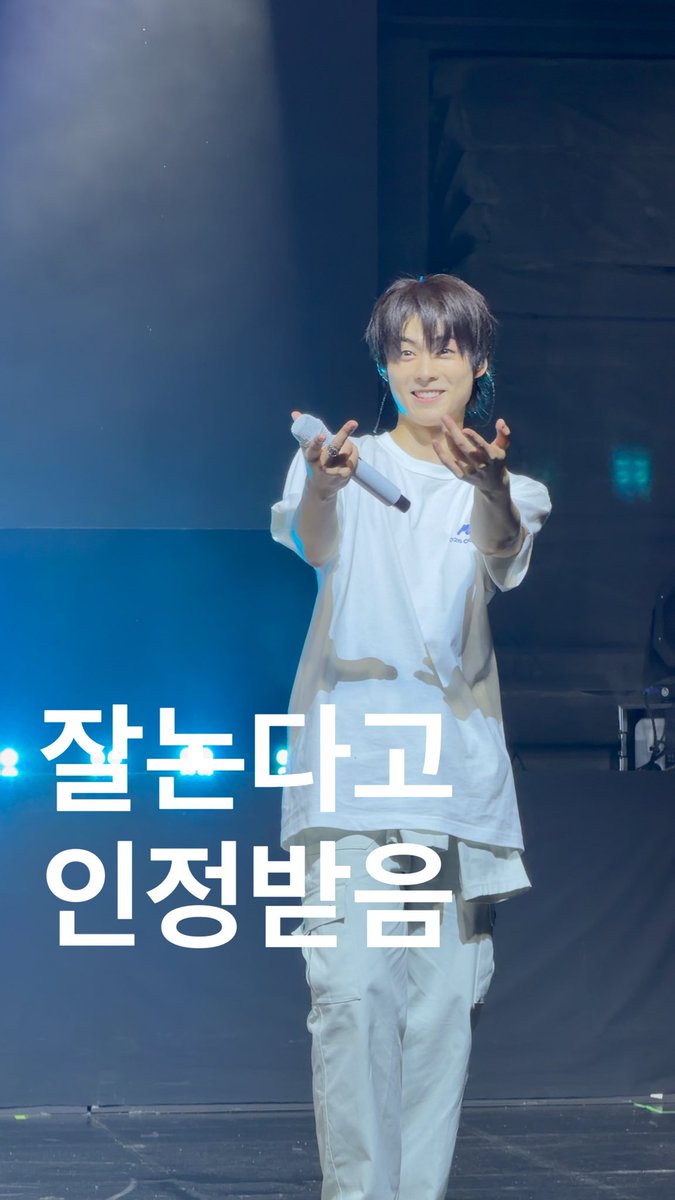 A young male performer on stage in a white t-shirt and pants holding a microphone, with Korean text "잘 놀다고 인정받음" (loosely: 'Acknowledged for having fun well') overlaid.