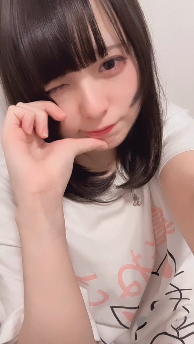 A close-up selfie of a young woman with dark hair winking at the camera while resting her chin on her hand, wearing a white graphic t-shirt.