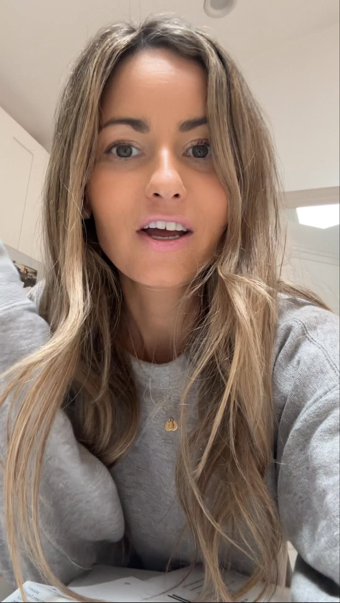 A woman with long blonde hair wearing a gray sweatshirt and a small gold pendant necklace appears to be speaking in a selfie-style video taken indoors.