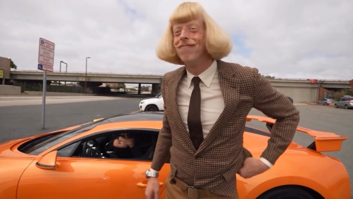 A man wearing a brown checkered blazer, white shirt, black tie, and a blonde wig with a mustache stands smiling next to an orange sports car in a parking lot.