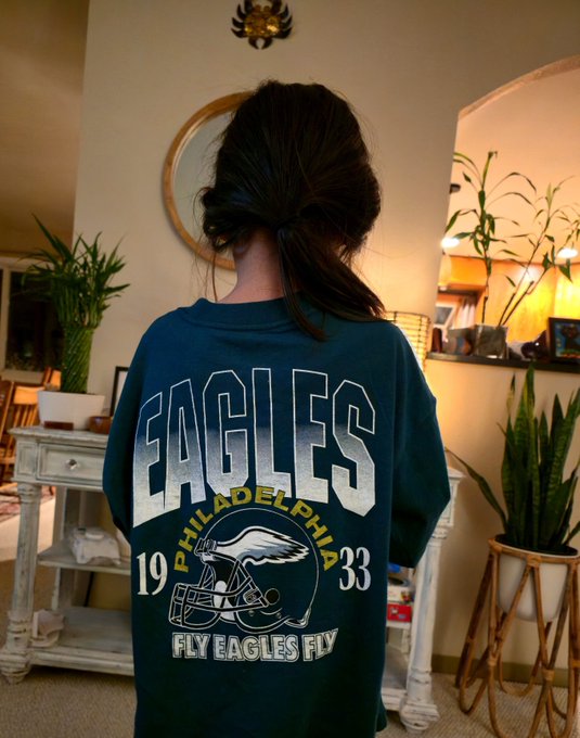 A person viewed from behind wears a green "EAGLES PHILADELPHIA 1933 FLY EAGLES FLY" jersey with a football helmet graphic in a room with plants and a round mirror.