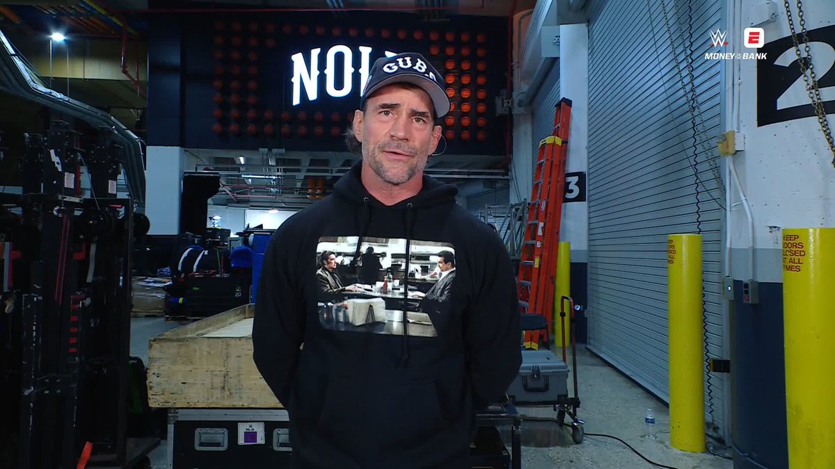 CM Punk, wearing a "CUBS" cap and a Pulp Fiction graphic hoodie, stands backstage in what appears to be an arena loading area, ahead of the Men's Money in the Bank Ladder Match being streamed on the WWE/ESPN platform.