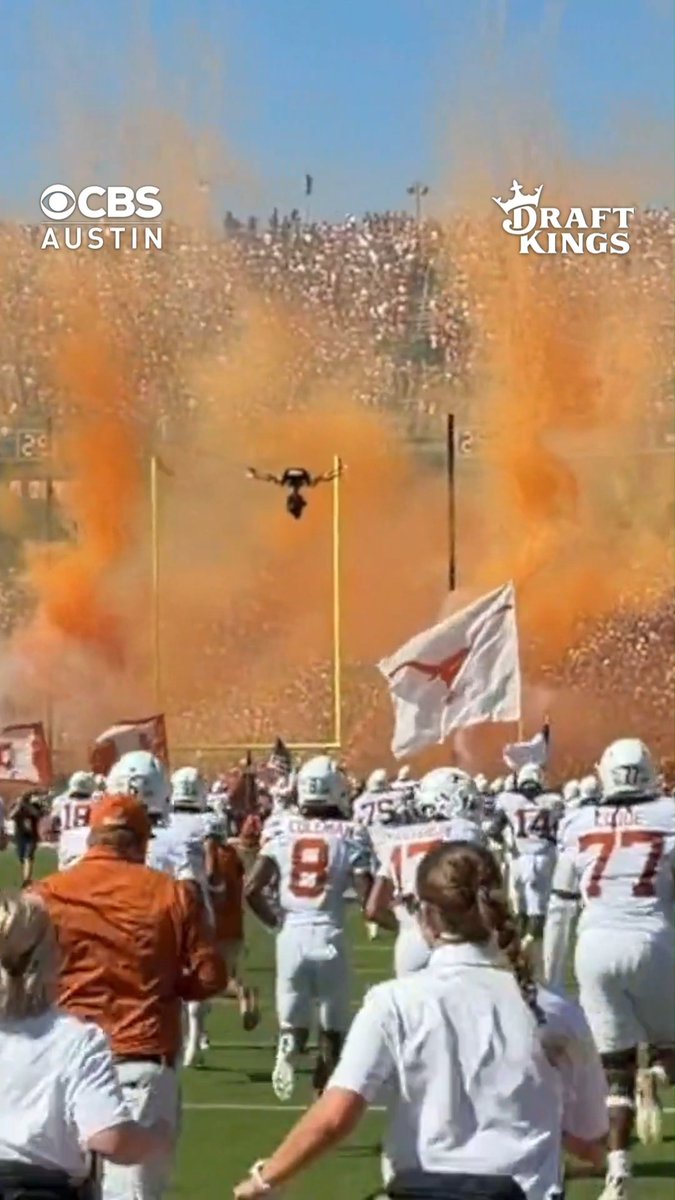 Texas football players in white jerseys (numbers 8 Coleman, 17, 77) charge onto the field through billowing orange smoke as Bevo, the Longhorns' mascot, leaps between the goalposts, while fans wave flags in a packed stadium—visually capturing the moment referenced in the CBS Austin/DraftKings broadcast about Texas leading Oklahoma 17-7 at halftime of the Red River Rivalry.