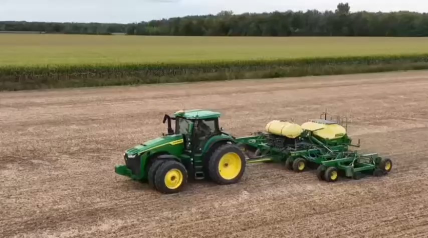 A green and yellow John Deere tractor pulling a planter is parked on a harvested agricultural field with trees in the background.