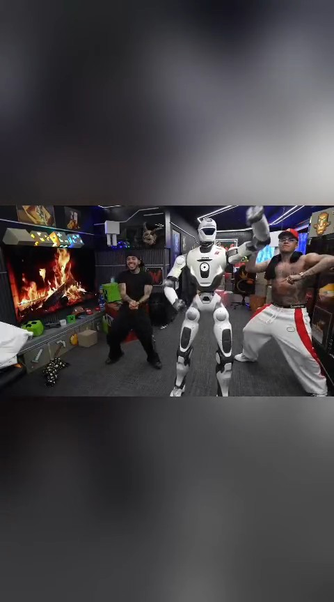 A scene from a game show or streaming set featuring a humanoid robot standing between two men, one wearing a black shirt and the other shirtless in a red cap and white pants, with a fireplace on a screen in the background.