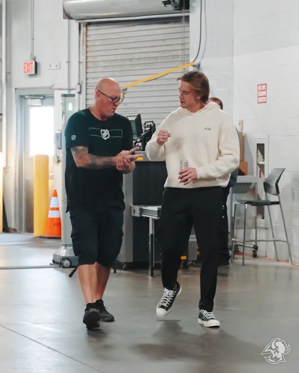 Two men converse in what appears to be a garage or warehouse setting, with a buffalo logo visible in the bottom right corner. The bald man on the left wears a black t-shirt with a logo and dark shorts, while the man on the right sports a cream-colored hoodie and black pants while holding a can. The "#LetsGoBuffalo" hashtag suggests this is a celebratory moment for a Buffalo team victory, possibly featuring Doaner, with the buffalo logo tying into the post's theme.
