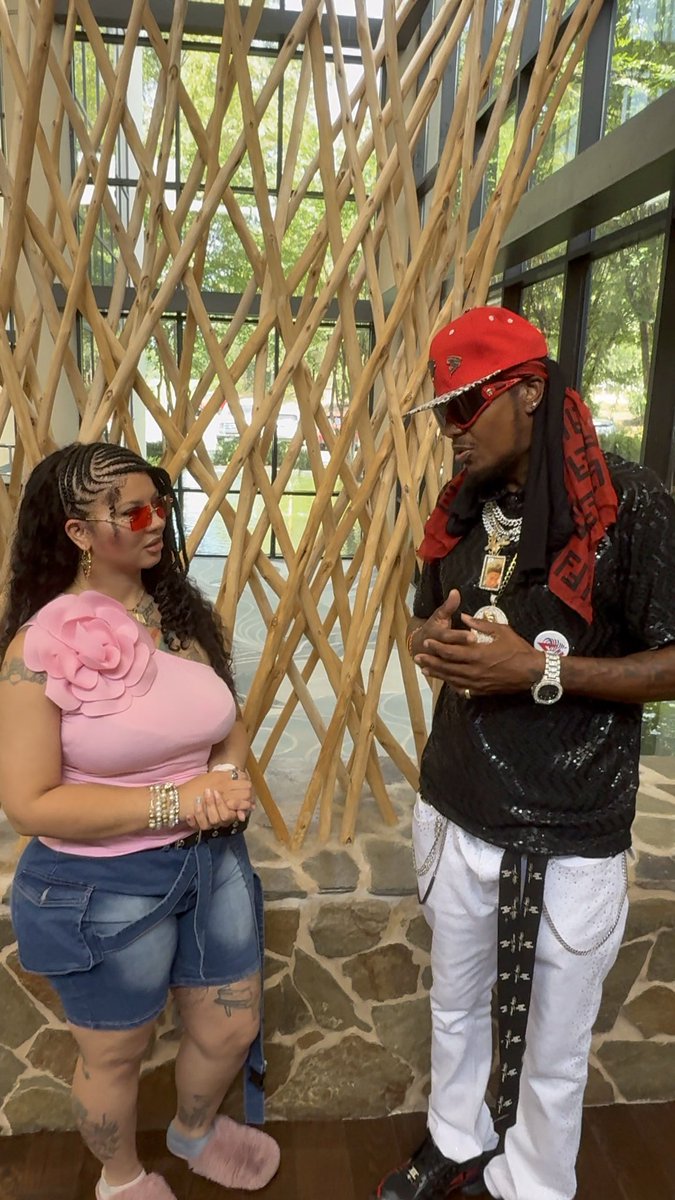 A woman wearing a pink one-shoulder top with a large flower detail and a denim skirt stands facing a man in a red cap, sunglasses, and black shirt, both adorned with jewelry, in front of a decorative branch installation.