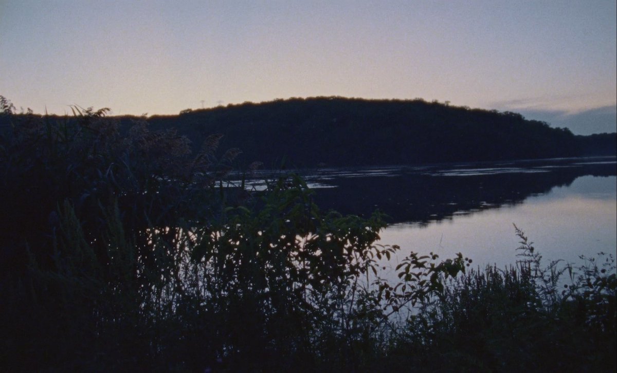 A scenic photograph taken at dusk showing a calm river reflecting the sky, with silhouetted hills, vegetation, and trees in the foreground.