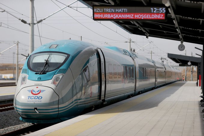 A sleek blue and white TCDD high-speed train stationed at a platform with an overhead sign reading "HIZLI TREN HATTI" and "IMHURBASKANIMIZ HOSGELDINIZ" showing 12:55.