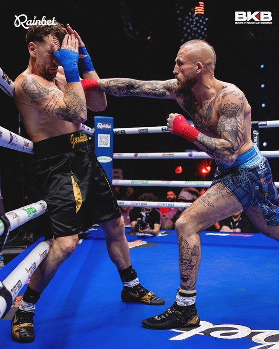 A bare-knuckle boxing match between two tattooed fighters in a ring sponsored by Rainbet and BKB (Bare Knuckle Boxing), with one fighter in black/gold trunks being punched by another in blue trunks.