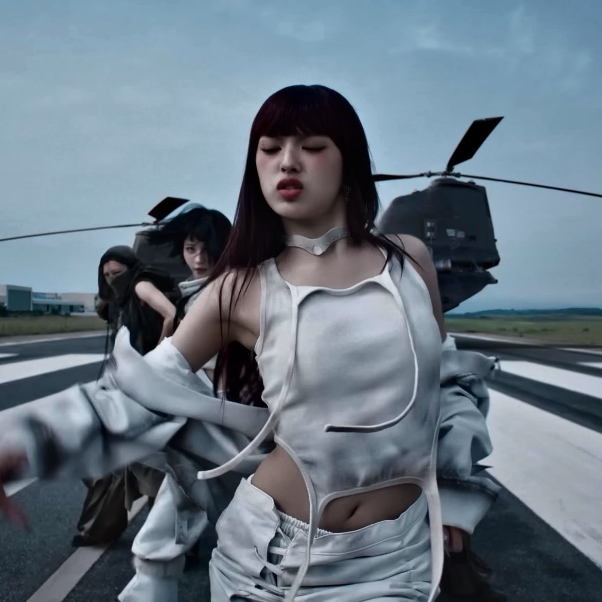 A cinematic shot of a young woman with long dark hair in a white outfit dancing on a runway with other figures and helicopters in the background.