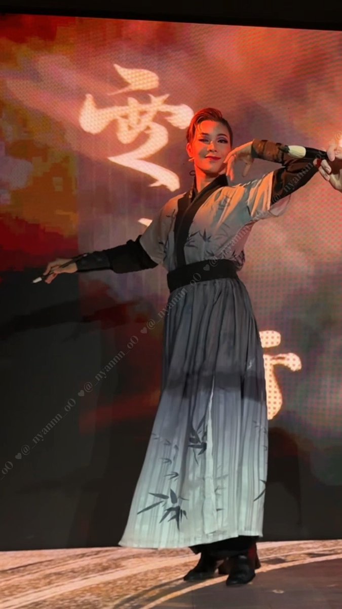 A woman with short hair performs on stage in a flowing white traditional-style outfit with bamboo patterns and a black sash, with Chinese characters "夢" (dream) projected behind her.