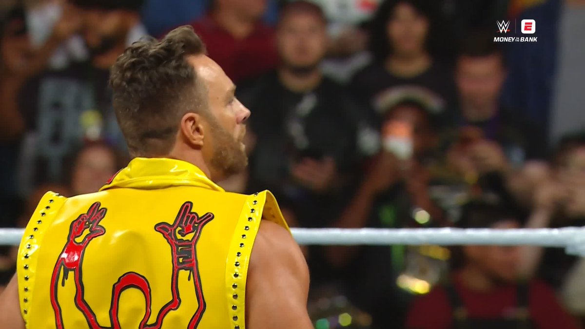A WWE wrestler with short brown hair and a beard stands in the ring wearing a vibrant yellow studded vest featuring a red dragon design on the back, looking out at a cheering crowd while the "Money in the Bank" logo appears in the upper right corner, capturing the electric atmosphere LA Knight is hushing the arena to hear.