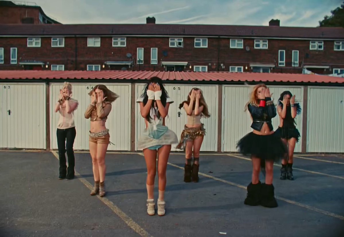 Group of seven women dancing in a parking lot in front of a row of garages and brick row houses, all covering their faces with their hands.