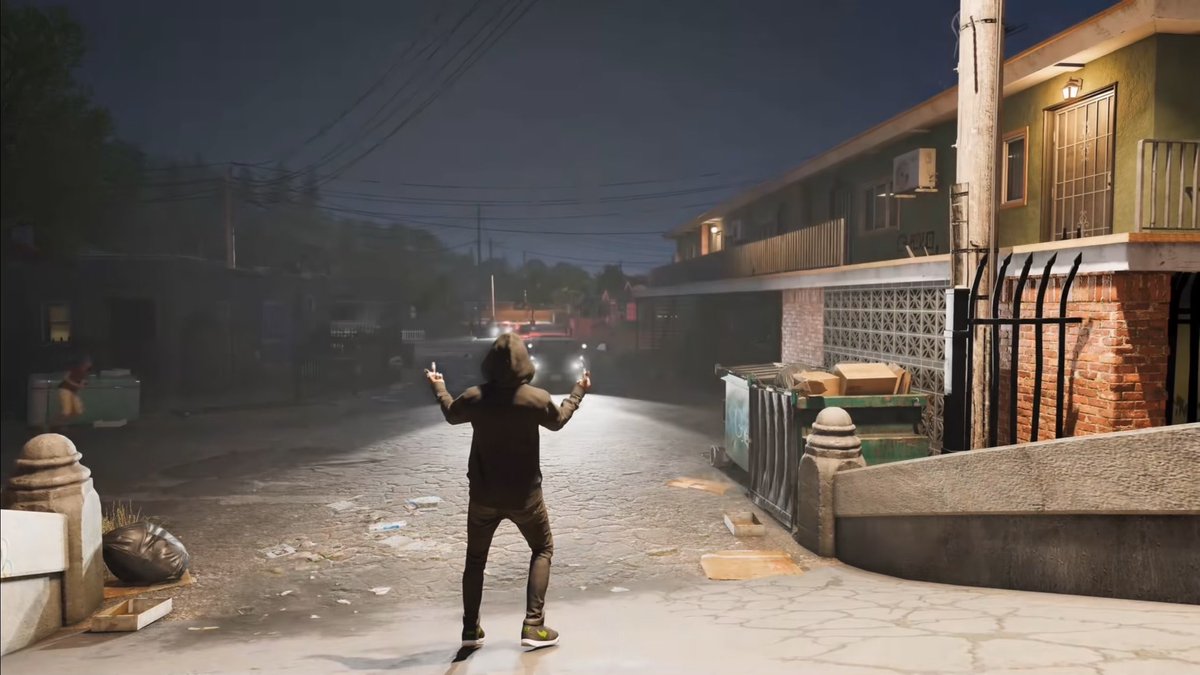 A person in a hooded sweatshirt stands in a dimly lit street, gesturing with both middle fingers raised toward approaching headlights, set in an urban environment with trash and dumpsters nearby.