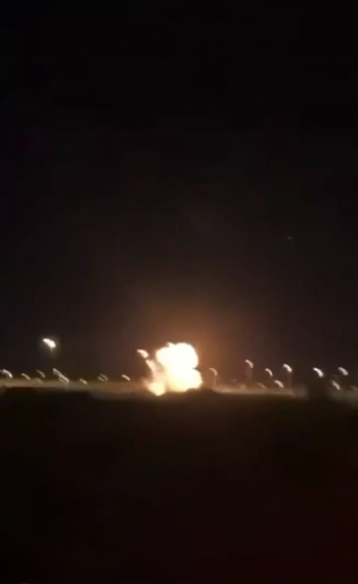 A nighttime photograph showing a bright explosion or fireball on the ground, with scattered lights visible in the dark surroundings.