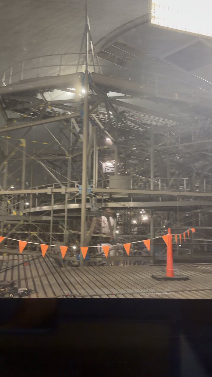 Interior view of a large dome-like structure under construction, showing extensive metal scaffolding and beams, with orange safety pennants and a traffic cone visible on a wooden platform in the foreground.