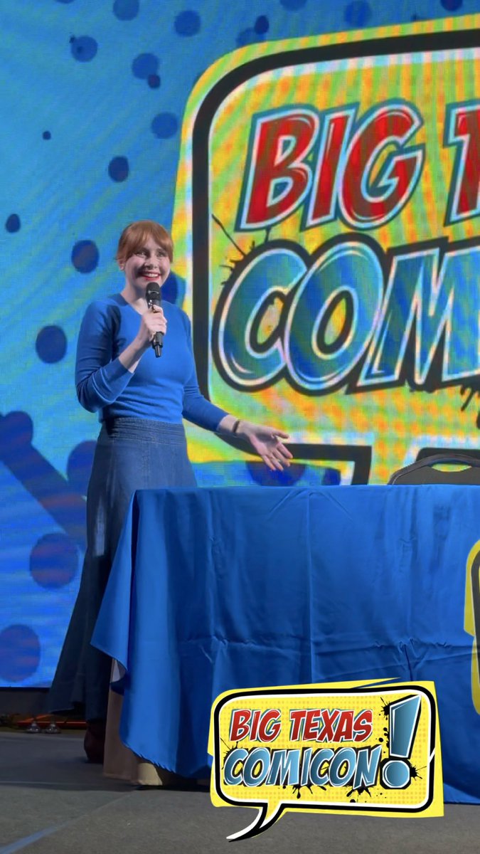 A woman with red hair in a blue top and denim skirt speaks into a microphone on stage at Big Texas Comicon, with the event's colorful logo visible behind her and on a table draped in blue.