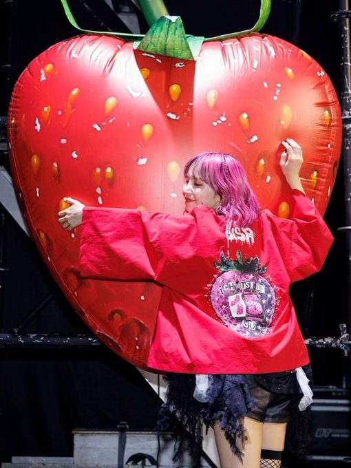 Stage photograph of a performer with pink hair wearing an oversized red jacket with 'FACE MISTER' text, standing in front of a giant inflatable strawberry prop.