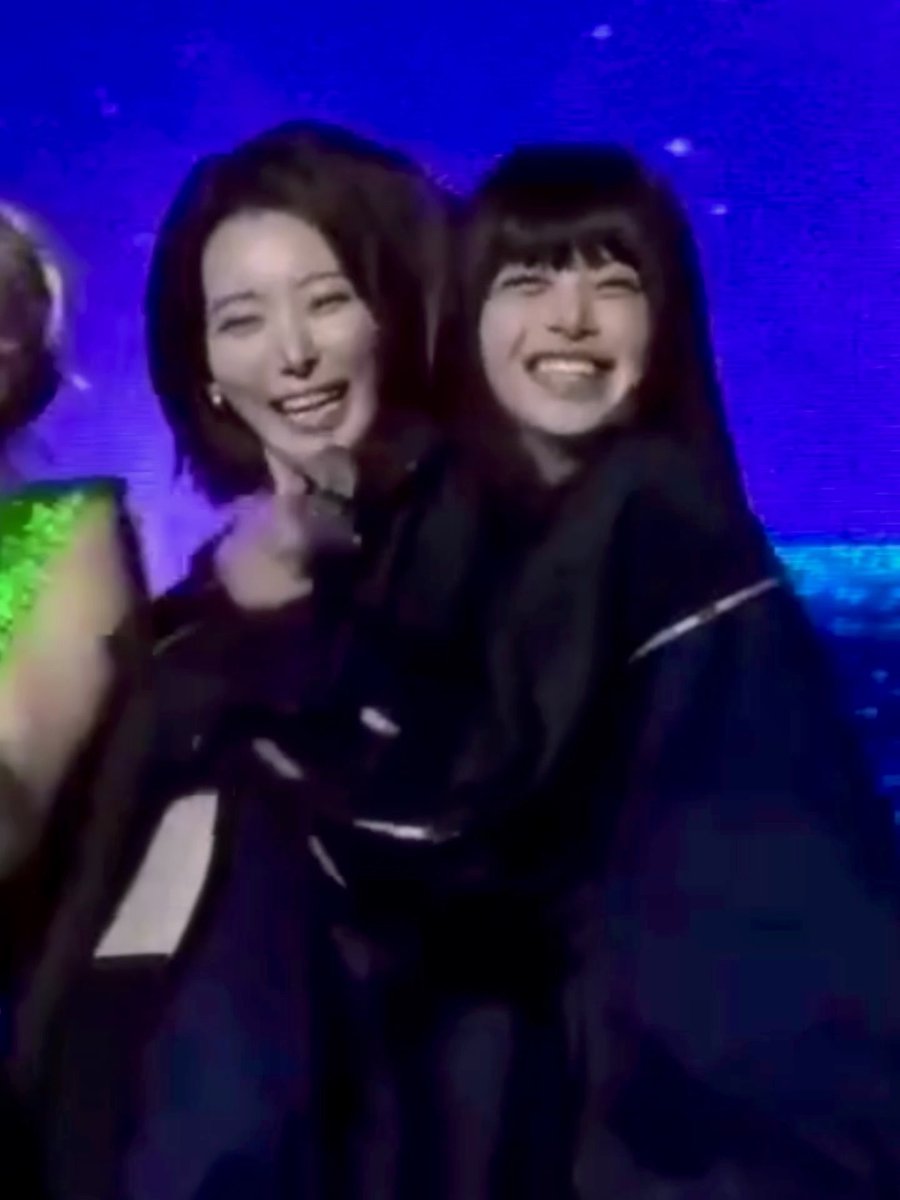 Two smiling young women stand together on a stage with a blue starry background, one wearing a dark outfit and the other in a dark jacket.