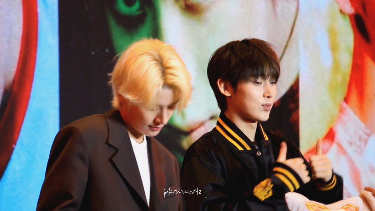 Two young men, one with blonde hair in a suit and one with dark hair in a varsity jacket giving thumbs up, pose in front of a large screen with a watermark 'jakehanart12'.