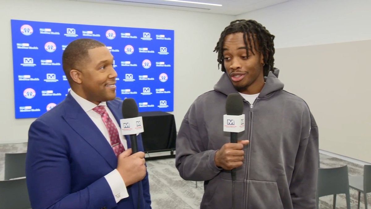 A sports reporter in a blue suit interviews a player wearing a grey hoodie, both holding microphones with a Monumental Sports logo, in front of a blue backdrop with team logos.