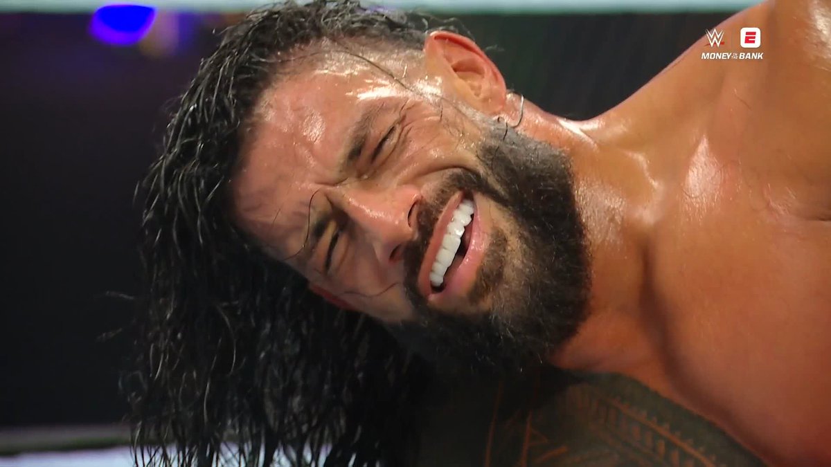 A close-up of a sweaty wrestler with long dark hair and a beard grimacing in pain during a WWE Money in the Bank match.