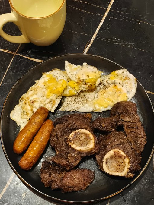 A hearty breakfast plate on a black dish contains fried eggs, two sausages, and braised beef shanks with bone marrow, accompanied by a yellow mug on a dark countertop.