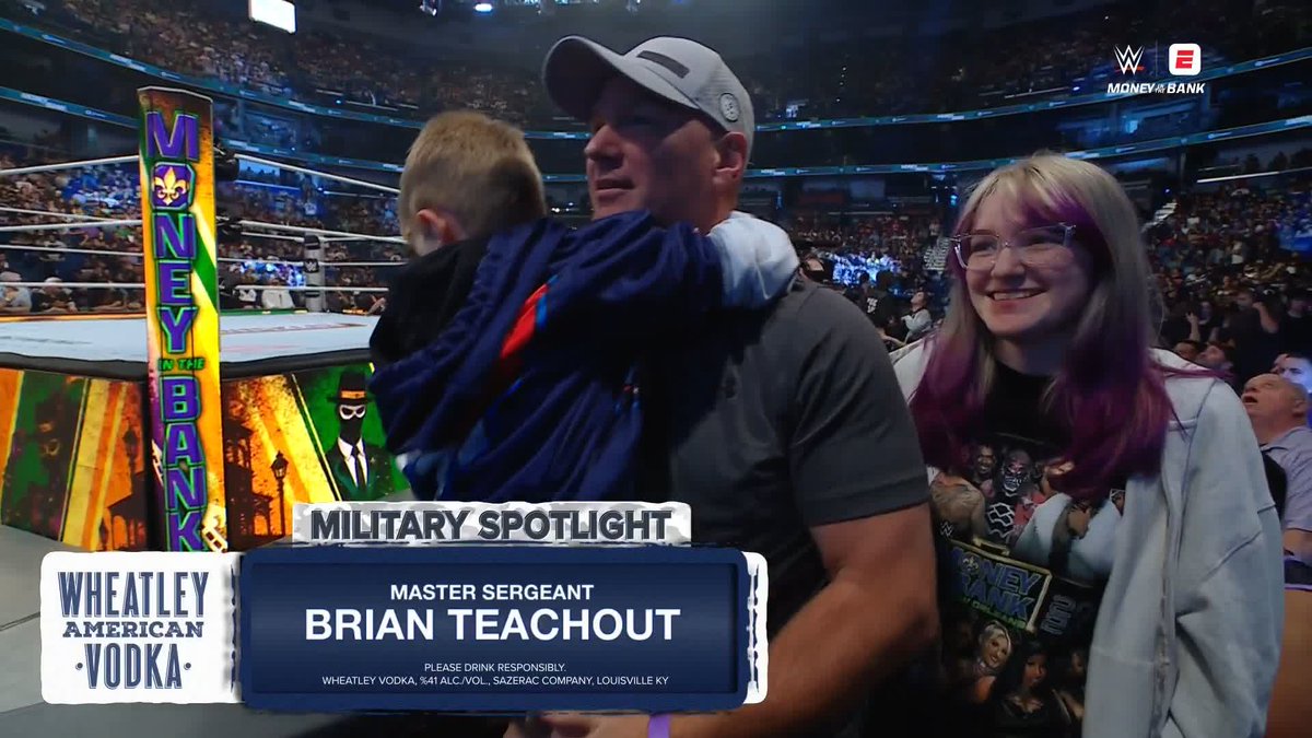 A man in a gray cap holding a young boy is shown alongside a smiling young woman with purple-streaked hair at a WWE Money in the Bank event, with on-screen text reading "MILITARY SPOTLIGHT: MASTER SERGEANT BRIAN TEACHOUT," presented by Wheatley American Vodka, honoring his service.