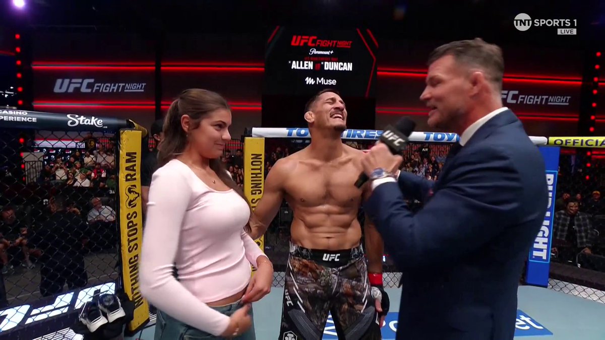 Niko Price stands in the Octagon with his daughter during a post-fight interview at UFC Vegas 122 (Allen vs. Duncan), laughing while a suited interviewer holds a UFC-branded microphone. The screen displays "TNT SPORTS 1 LIVE," "UFC FIGHT NIGHT," and sponsor signage for Stake and Toyo Tires.