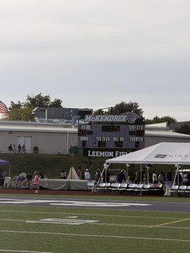 A view of a football field with white sideline tents set up along the edge, where a scoreboard in the background displays 'LACKAWANNA' and 'LEMON FIELD' alongside an American flag.