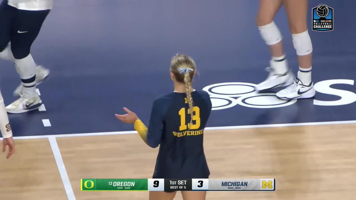 A volleyball player wearing a navy Michigan Wolverines jersey with the number 13 stands on the court during a match against Oregon, with the scoreboard showing Oregon 9, Michigan 3 in the 1st set of a Best of 5.