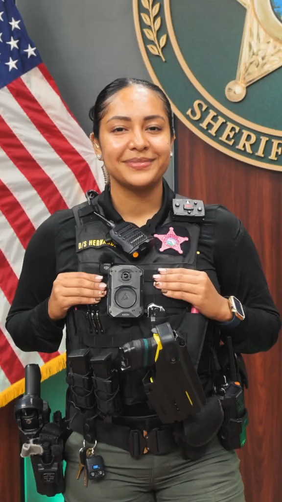 A smiling female sheriff's deputy in a black uniform with badge stands in front of an American flag and a sheriff's department seal, with her duty belt visible.