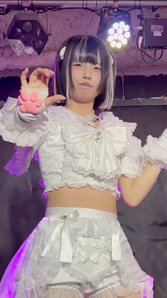 A young woman with short dark hair styled in a bob, wearing an elaborate white frilly dress with ribbons and bows, posing at what appears to be a concert or stage venue with stage lighting visible overhead.
