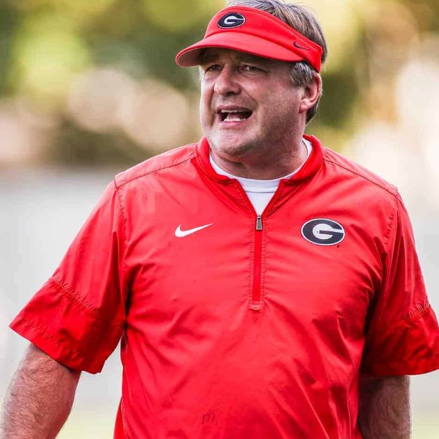 A close-up photograph of Georgia Bulldogs football coach wearing a red Nike quarter-zip jacket and red visor with the 'G' logo, appearing to shout instructions during a practice or game.