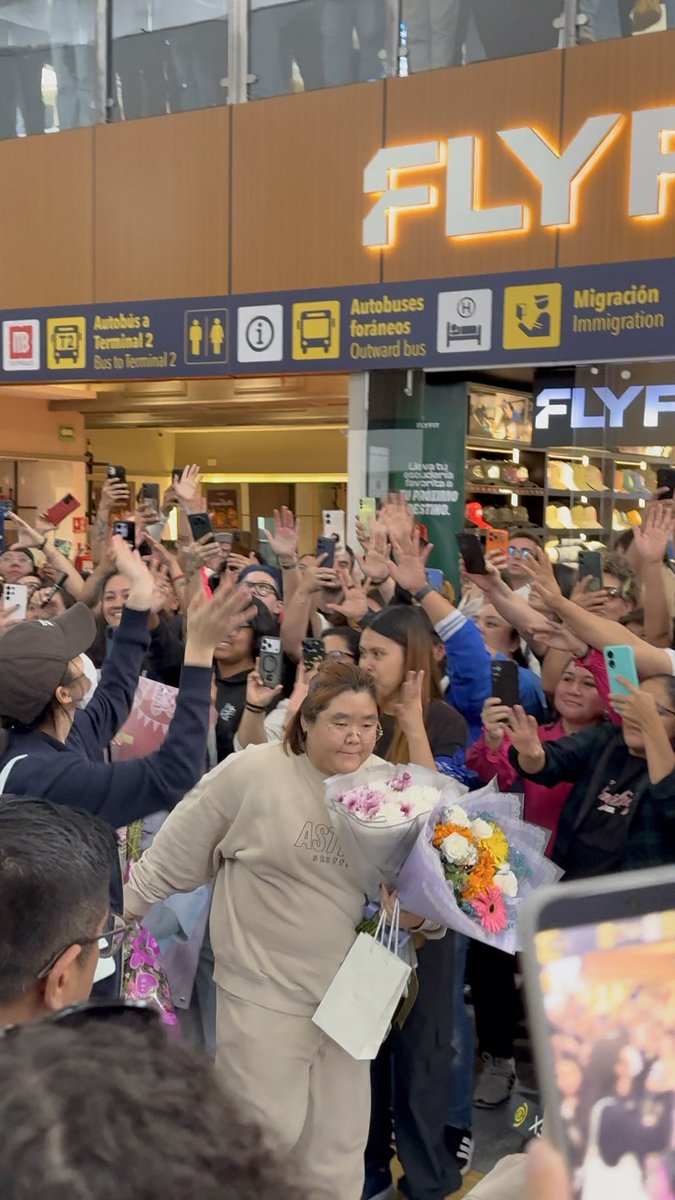 A crowd of people holding up phones to greet a woman carrying a bouquet of flowers at an airport, with signs for 'FLY' and 'Migración Immigration' visible.