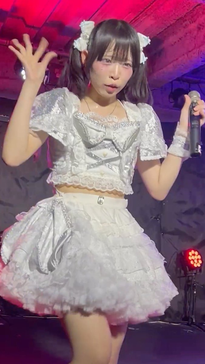 A female J-pop performer in a silver sparkly two-stage outfit with pigtails and white bows, holding a microphone on stage.