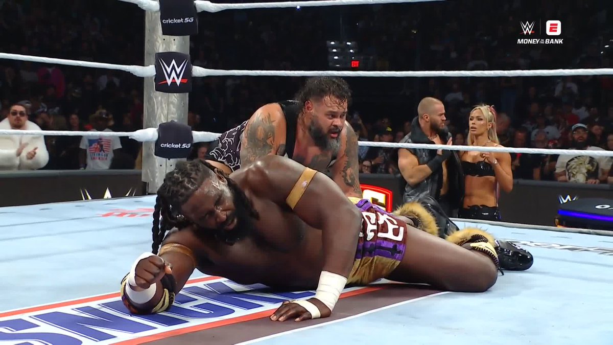 A WWE wrestling match scene from Money in the Bank, showing two male wrestlers down on the mat while another pair stands in the ring's corner in front of a cheering crowd.