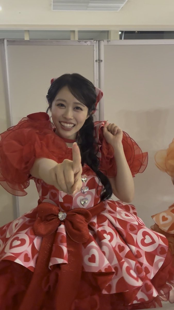 A smiling young woman in a vibrant red dress with heart patterns poses with a thumbs-up gesture.